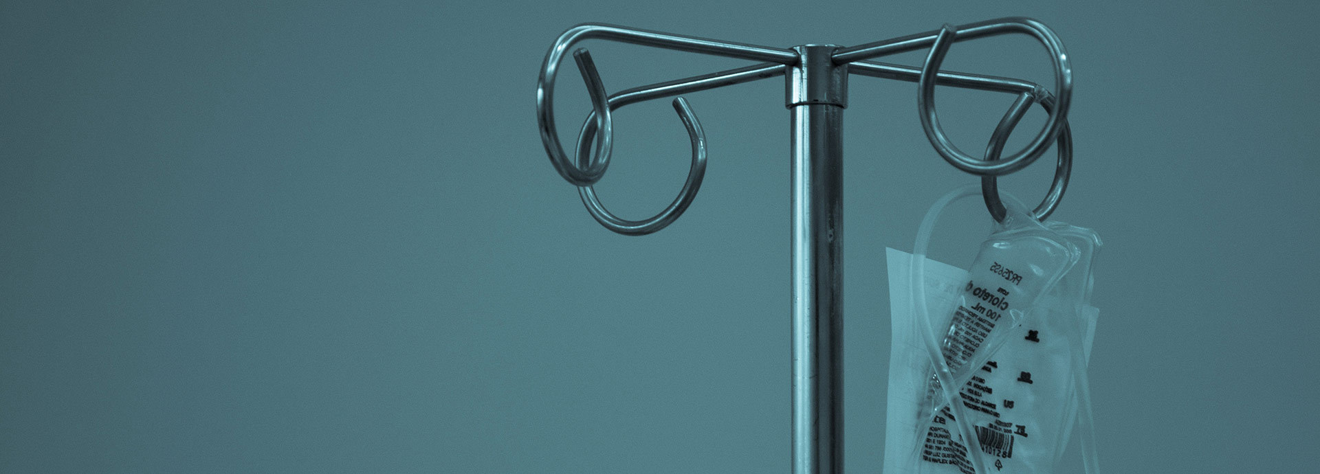 A metal IV stand with coiled hooks holds a clear, labeled medical fluid bag against a muted blue-gray background, conveying a clinical and sterile atmosphere.